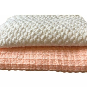 Peut inclure: Deux couvertures pour bébé en tricot, une blanche et une pêche. Les deux couvertures ont un motif texturé et tissé.