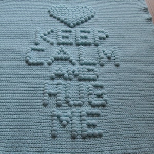 May include: A light blue crocheted baby blanket with the words "Keep Calm and Hug Me" in raised white stitches. The blanket features a heart at the top.