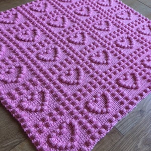 May include: A pink crocheted baby blanket with a heart pattern. The blanket is made with a textured stitch that creates a raised effect.