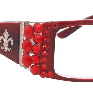May include: Burgundy-coloured eyeglasses with a rectangular frame. The temples feature a silver fleur-de-lis and red jewel embellishments. The frame is decorated with red jewels on the sides.