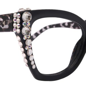 Könnte beinhalten: Schwarze Brille im Cat-Eye-Stil mit Leopardenmuster auf den Bügeln. Die Fassungen sind mit klaren Strasssteinen verziert. Die Brille hat ein auffälliges, glamouröses Design.