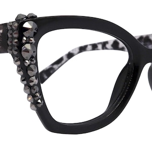 Könnte beinhalten: Schwarze Cat-Eye-Brille mit Leopardenmuster an den Bügeln. Die Fassungen sind mit dunkelgrauen Strasssteinen verziert. Die Brille hat ein auffälliges, stilvolles Design.