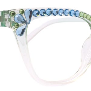 Peut inclure: Lunettes à monture transparente ornées de strass bleus et verts. Les montures œil de chat présentent un motif floral sur les coins supérieurs et une rangée de strass sur le bord supérieur. Les branches sont de couleur vert clair.