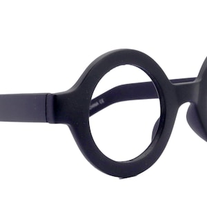 Op de afbeelding: Zwarte ronde bril met dikke monturen en rechte pootjes. De bril heeft een matte afwerking en een klassiek, minimalistisch design. De glazen zijn helder en de neuspads zijn in het montuur geïntegreerd.
