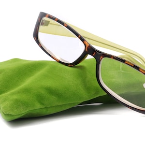 Könnte beinhalten: Lesebrille mit einem Rahmen im Schildpattmuster und grünen Bügeln liegt auf einem grünen Samtbeutel. Das rechte Glas hat eine Vergrößerung von +6,00. Die Brille ist zum Lesen konzipiert und wird mit einem Etui geliefert.