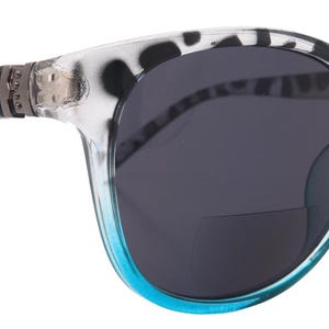 Puede incluir: Gafas de sol con montura transparente y estampado de lunares en blanco y negro en las patillas. Las lentes son de color gris oscuro y la parte inferior de la montura es azul turquesa. Diseño de ojo de gato.