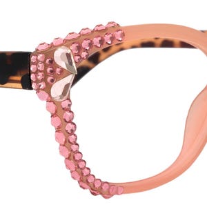 Könnte beinhalten: Brille im Cat-Eye-Stil mit pfirsichfarbenem Rahmen und Bügeln mit Leopardenmuster. Der Rahmen ist mit rosa Strasssteinen und einem herzförmigen Akzent verziert. Ein modisches Accessoire.