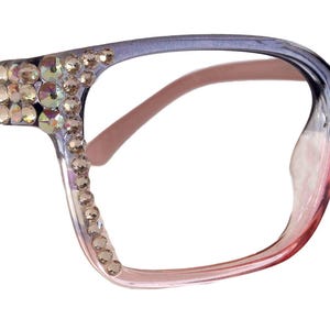 Può includere: Un paio di occhiali blu e rosa con strass sulla montatura. Gli occhiali hanno una forma quadrata e lenti trasparenti.