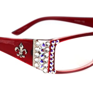 Może przedstawiać: Okulary plastikowe w kolorze czerwonym z przezroczystymi szkłami i rzędem cyrkonii po bokach. Oprawka ma srebrny wzór lilii.