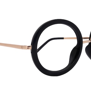 Könnte beinhalten: Schwarze Brille mit rundem Rahmen, goldenen Bügeln und schwarzen Ohrenstücken. Die Brille hat ein dickes, auffälliges Rahmendesign. Die Gläser sind klar. Die Brille ist vor einem weißen Hintergrund platziert.