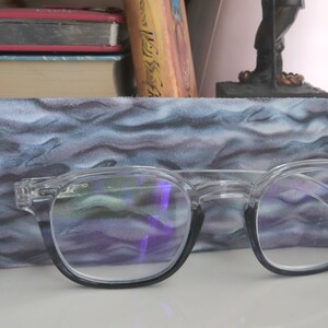Op de afbeelding: Een bril met heldere glazen en zwarte monturen die op een blauwe en grijze stoffen etui met patroon ligt. De bril heeft een lichte blauwe tint in de glazen.