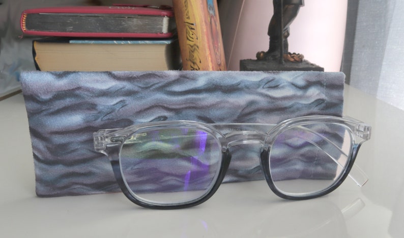 Op de afbeelding: Een bril met heldere glazen en zwarte monturen die op een blauwe en grijze stoffen etui met patroon ligt. De bril heeft een lichte blauwe tint in de glazen.