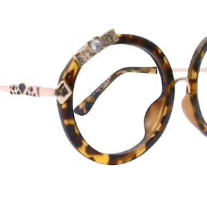 Puede incluir: Gafas redondas de carey con patillas de metal dorado y detalles decorativos. Las patillas tienen un diseño en forma de corazón y un pequeño adorno de cristal.