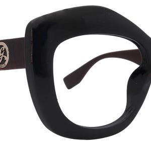 May include: Black oversized eyeglasses with a bold, angular frame design. The temples feature a reddish-brown woodgrain pattern and a silver logo. The lenses are clear, and the overall style is retro-inspired.