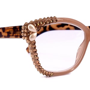 Könnte beinhalten: Eine beige Cat-Eye-Brille mit klaren Gläsern. Die Bügel sind mit Strasssteinen und einem perlmuttartigen Schmuckstück auf jeder Seite verziert.