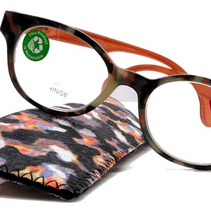 May include: Reading glasses with a tortoiseshell pattern frame and orange accents. The glasses have a +2.75 magnification and are resting on a patterned case. The frame has the word "HINGE" printed on it and a "This Product is Recyclable" sticker.