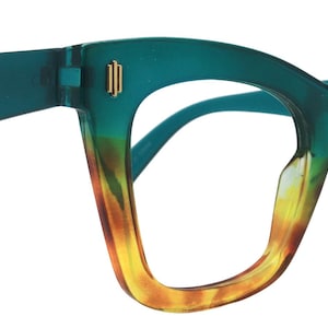 Puede incluir: Un par de gafas degradadas en verde azulado y amarillo. Las monturas son gruesas y angulares, con un degradado que va del verde azulado al amarillo. Las patillas son verde azulado y las lentes son transparentes. Acentos dorados en la montura.