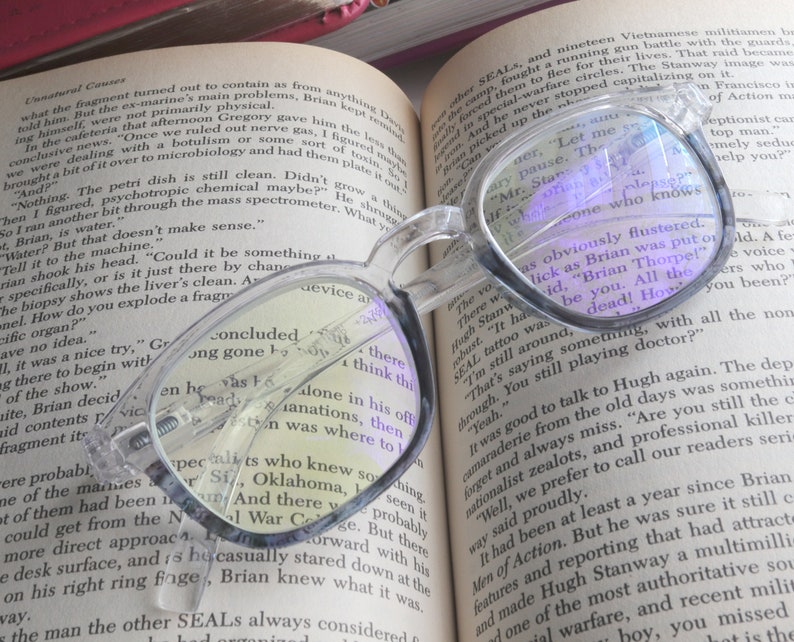Op de afbeelding: Een close-up van een helder paar brillen dat op een open boek ligt. De bril heeft een blauwe tint en bedekt gedeeltelijk de tekst op de pagina. De tekst op de pagina gaat over een militaire inval en de nasleep van een gevecht.