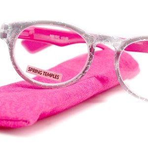 May include: Clear reading glasses with a sparkly silver frame and pink spring hinges. The glasses are in a pink case and have a label that says "Spring Temples". The glasses have a +3.00 lens strength.
