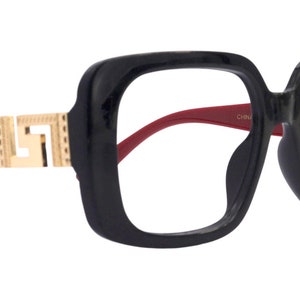 Könnte beinhalten: Schwarze quadratische Brille mit weinroten Bügeln und goldfarbenen Zierakzenten. Die Brille hat ein kühnes, modernes Design. Die Gläser sind klar.