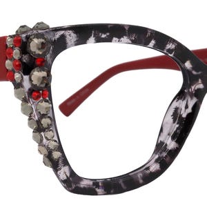 Könnte beinhalten: Brille im Cat-Eye-Stil mit rotem Gestell und Bügeln. Die Gläser sind von einem schwarz-weißen Fleckenmuster umgeben. Die Bügel sind mit roten und silbernen Strasssteinen und Nieten verziert. Die Brille hat ein auffälliges, modisches Design.