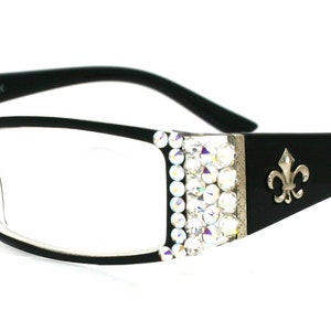May include: Black rectangular reading glasses with clear lenses. The frames have a decorative silver accent with clear rhinestones on the sides and a fleur-de-lis emblem on the temples. The glasses are set against a white background.