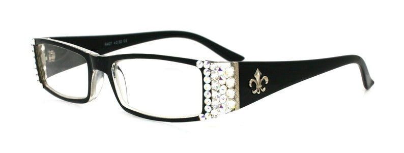 May include: Black rectangular reading glasses with clear lenses. The frames have a decorative silver accent with clear rhinestones on the sides and a fleur-de-lis emblem on the temples. The glasses are set against a white background.