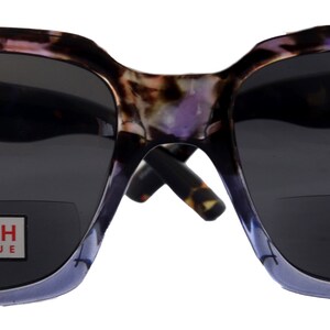May include: A pair of tortoiseshell and purple sunglasses with a blue frame and grey lenses. The sunglasses have a reading glass insert with the text "NY FIFTH AVENUE" printed on it.
