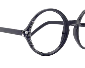Puede incluir: Gafas negras de montura redonda con adornos decorativos. Las monturas son de color negro mate, con pequeñas piedras oscuras que acentúan los bordes exteriores de las lentes. Las varillas son finas y rectas.