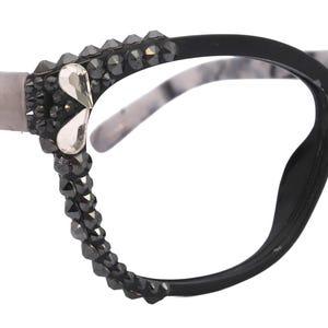 Könnte beinhalten: Schwarze Cat-Eye-Brille mit Bügeln mit grau-weißem Marmormuster. Die Fassungen sind mit dunkelgrauen Kristallen und einem silberfarbenen Akzent verziert. Die Brille hat ein stilvolles, modernes Design.