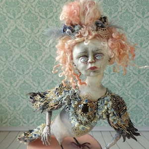 Könnte beinhalten: Eine handgefertigte Puppe mit einem weißen Gesicht, rosafarbenen lockigen Haaren und einem blauen und goldenen Federkostüm. Die Puppe hat große, weiße Augen und einen ernsten Ausdruck.