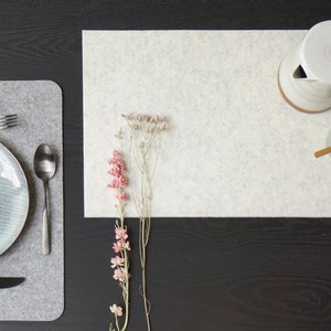 May include: A gray felt placemat with a blue and white striped plate, a fork, two spoons, and a knife. The placemat is on a black wooden table. There is a white felt placemat with dried flowers on the table.
