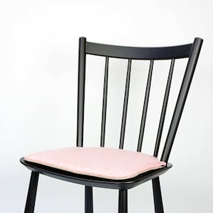 May include: A black wooden chair with a spindle back and a square seat. The seat has a light pink cushion. The chair has four legs and a crossbar for support. The chair is set against a white background.