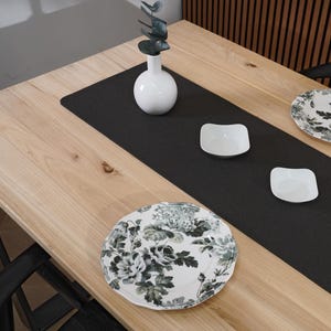 May include: A wooden dining table with a black runner, white dishes, and floral patterned plates. A white vase with greenery sits on the table. The table is set for a meal.