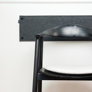 May include: Two black wooden chairs with white seat cushions are positioned against a white wall. A dark gray rectangular panel is mounted on the wall above the chairs. A potted plant on a light wood stool is on the left.