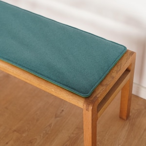 Puede incluir: Un banco de madera con un coj&iacute;n verde azulado. El banco tiene un coj&iacute;n rectangular verde azulado con un borde verde. El marco de madera es de color marr&oacute;n claro. El banco est&aacute; sobre un suelo de madera.