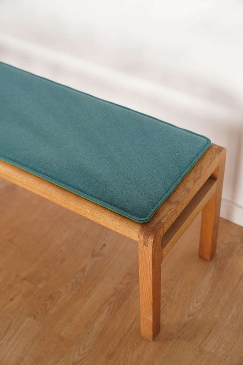 Puede incluir: Un banco de madera con un coj&iacute;n verde azulado. El banco tiene un coj&iacute;n rectangular verde azulado con un borde verde. El marco de madera es de color marr&oacute;n claro. El banco est&aacute; sobre un suelo de madera.