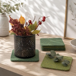 May include: A decorative arrangement featuring a black metal perforated vase holding fall foliage with red berries. Square green felt coasters are arranged around the vase, with a stack of coasters in the background. The scene is set on a light wood surface.
