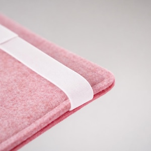 May include: Close-up of a pink felt sleeve with a white elastic band. The sleeve has a rounded corner and a textured surface. The white band is positioned diagonally across the sleeve.