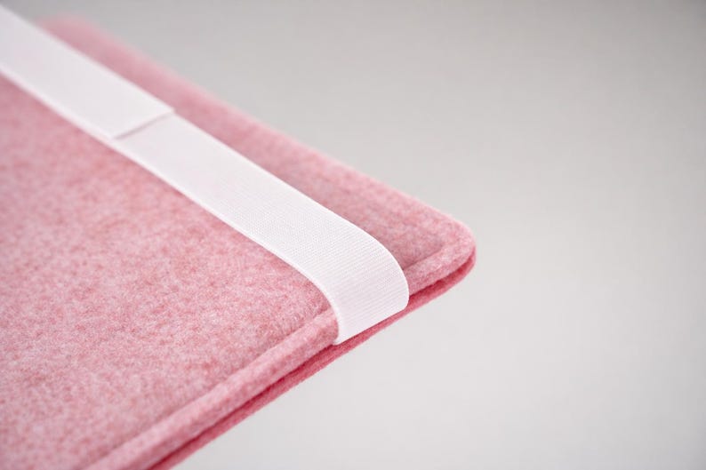 May include: Close-up of a pink felt sleeve with a white elastic band. The sleeve has a rounded corner and a textured surface. The white band is positioned diagonally across the sleeve.