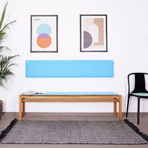 K&ouml;nnte beinhalten: Ein modernes Interieur mit einer Holzbank und einem hellblauen Kissen, einem schwarzen Stuhl und einem grauen Teppich mit Quasten. Zwei gerahmte abstrakte Kunstwerke h&auml;ngen &uuml;ber der Bank, und eine gro&szlig;e Topfpflanze f&uuml;gt Gr&uuml;n hinzu.