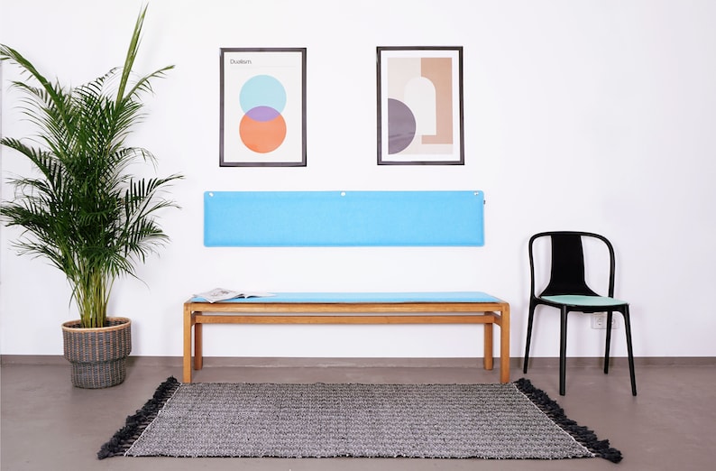 K&ouml;nnte beinhalten: Ein modernes Interieur mit einer Holzbank und einem hellblauen Kissen, einem schwarzen Stuhl und einem grauen Teppich mit Quasten. Zwei gerahmte abstrakte Kunstwerke h&auml;ngen &uuml;ber der Bank, und eine gro&szlig;e Topfpflanze f&uuml;gt Gr&uuml;n hinzu.