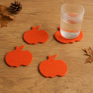 Könnte beinhalten: Ein Set aus vier orangefarbenen Filzuntersetzern in Kürbisform, wobei ein klares, geripptes Glas auf einem der Untersetzer steht. Die Untersetzer sind flach, kreisförmig mit einem kleinen Stielansatz und haben eine weiche, matte Textur.