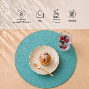 Könnte beinhalten: Ein türkisfarbener runder Tischset aus recycelten PET-Flaschen. Ein Teller mit einem Muffin und einer goldenen Gabel liegt auf dem Tischset. Ein Glas Wasser mit Beeren steht neben dem Teller. Die Worte "cozy", "made in Cologne" und "washable" sind enthalten.