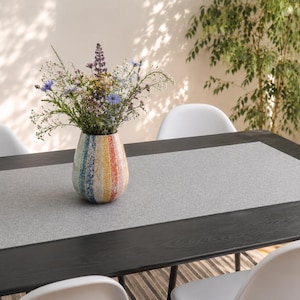 May include: A dining table set with a gray table runner and a colorful vase filled with flowers. The vase has a rainbow design. White chairs surround the dark wood table, and a plant is in the background.