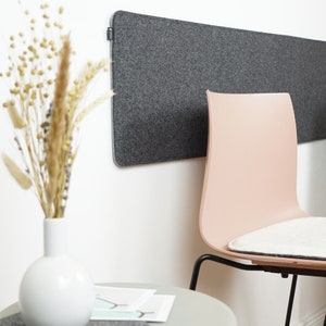 Op de afbeelding: Een grijs vilt geluiddempend paneel is aan een witte muur bevestigd. Een lichtroze stoel met een zwart metalen frame staat eronder. Een witte vaas met gedroogde bloemen en een kleine grijze tafel maken de scène compleet.