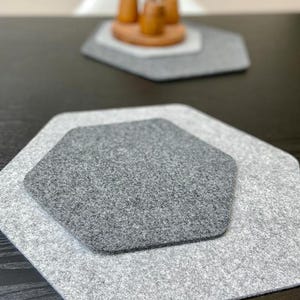 Può includere: Un set di tre tovagliette in feltro grigio a forma esagonale. Le tovagliette sono in diverse tonalità di grigio, con un tappetino grigio scuro nidificato all'interno di un tappetino grigio chiaro. I tappetini sono su una superficie scura.