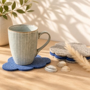 May include: A ceramic mug with vertical white stripes sits on a blue cloud-shaped coaster. Additional cloud-shaped coasters in gray and blue are stacked nearby. Small seashells and a vase with greenery complete the scene.