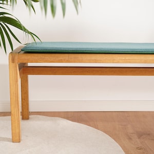 Puede incluir: Un banco de madera con un coj&iacute;n verde azulado. El banco tiene forma rectangular con cuatro patas y una viga de soporte inferior. El banco es de madera clara y est&aacute; colocado frente a una pared blanca. Una planta en una maceta blanca es visible a la izquierda.