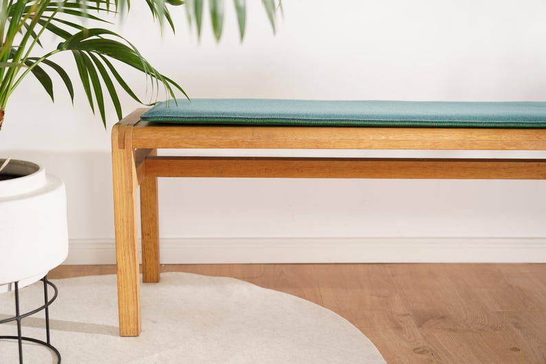 Puede incluir: Un banco de madera con un coj&iacute;n verde azulado. El banco tiene forma rectangular con cuatro patas y una viga de soporte inferior. El banco es de madera clara y est&aacute; colocado frente a una pared blanca. Una planta en una maceta blanca es visible a la izquierda.